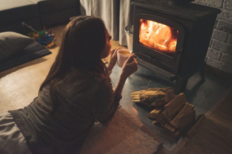 A woman drinks a mug of cocoa watching a fire burn in a wood-stove. We Can GIve You Safer Woodstove Home Heating Done Right!